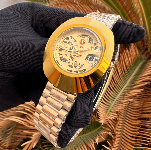 Gold wristwatch with a black strap held by a gloved hand against a natural background
